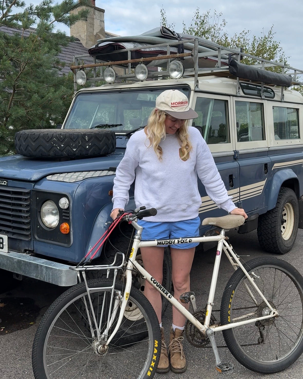 A woman stands outdoors in front of a blue vintage Land Rover with a roof rack, holding a white Commencement bicycle. She is wearing a white sweatshirt, blue shorts, and brown boots, with a surfboard on the vehicle roof.