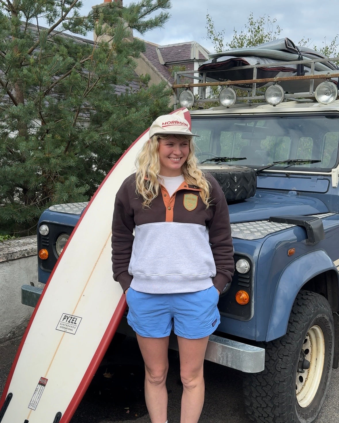 A woman stands in front of a blue vintage Land Rover with a surfboard leaning against it. She is wearing a brown, white, and orange color-blocked quarter-zip sweatshirt, blue shorts, and a white cap with red text.