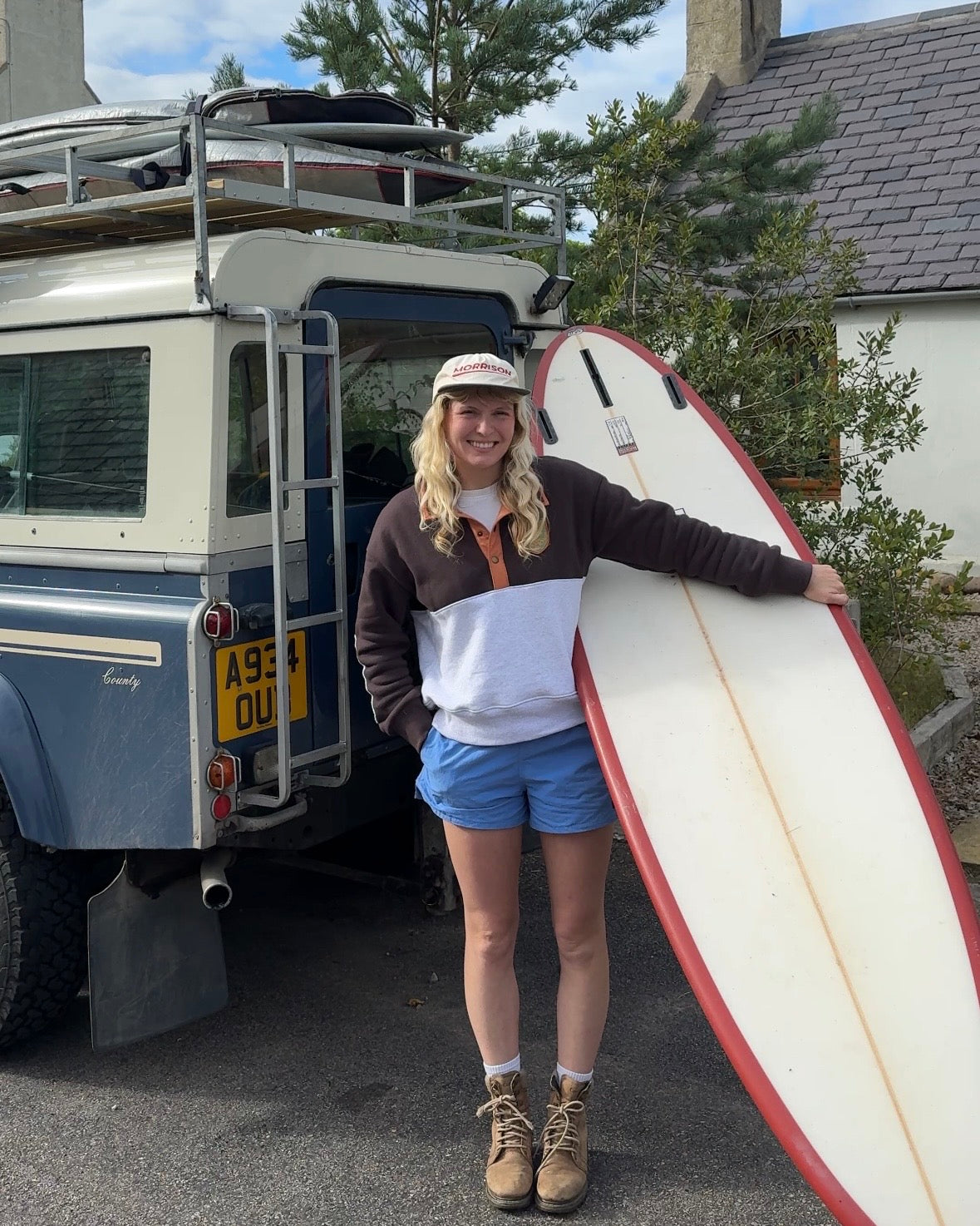 A woman stands outdoors holding a long surfboard next to a vintage camper van. She is wearing a colorblock sweatshirt, blue shorts, and hiking boots, with a house and trees in the background.