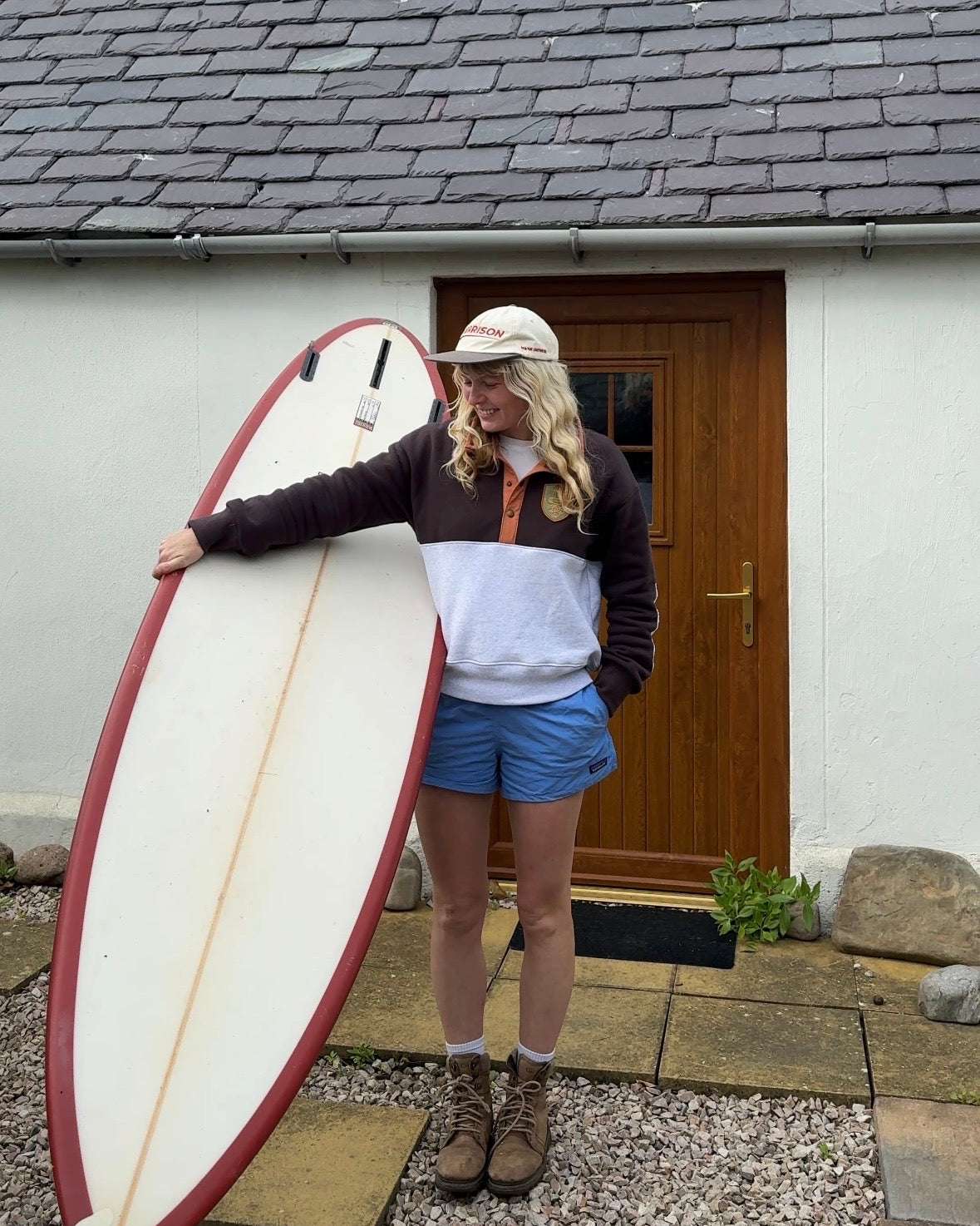 A woman stands outdoors in front of a white building with a wooden door, holding a large surfboard. She is wearing a colorblock pullover with a dark brown and white body, blue shorts, brown boots, and a beige cap.
