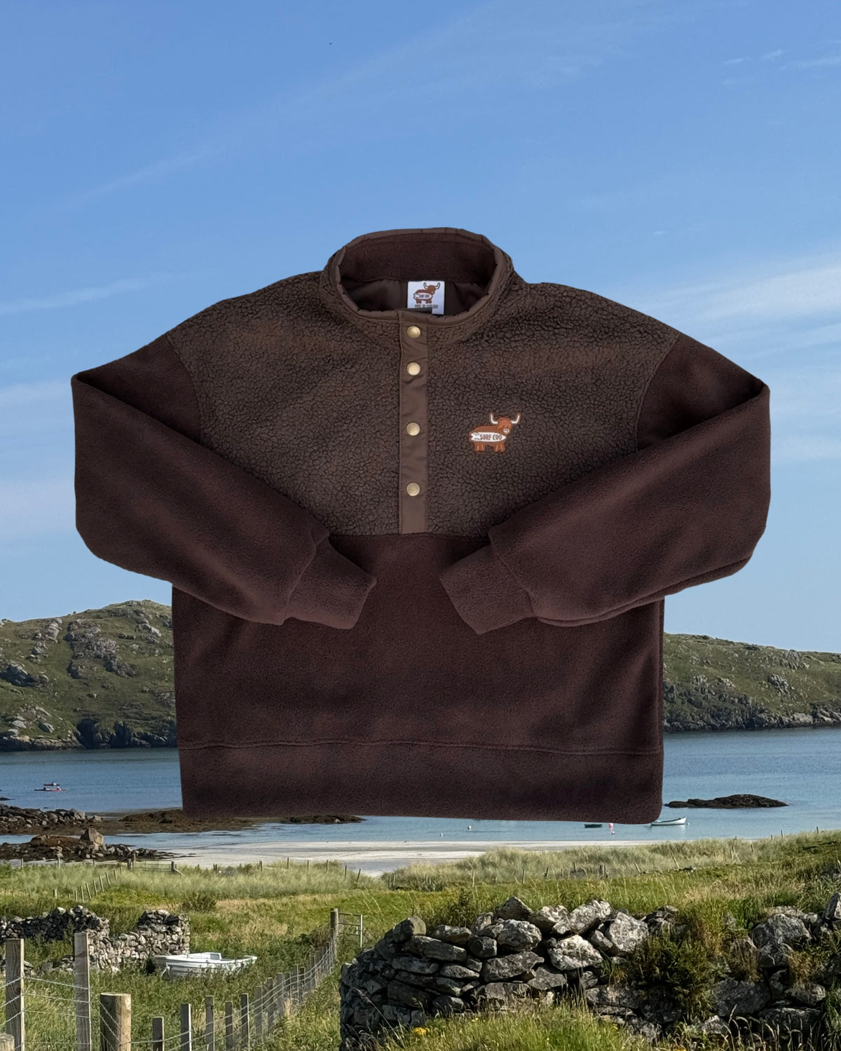 A brown fleece pullover sweater with a buttoned collar and a small deer logo on the chest, shown against a coastal landscape background with water, hills, and grassy foreground.