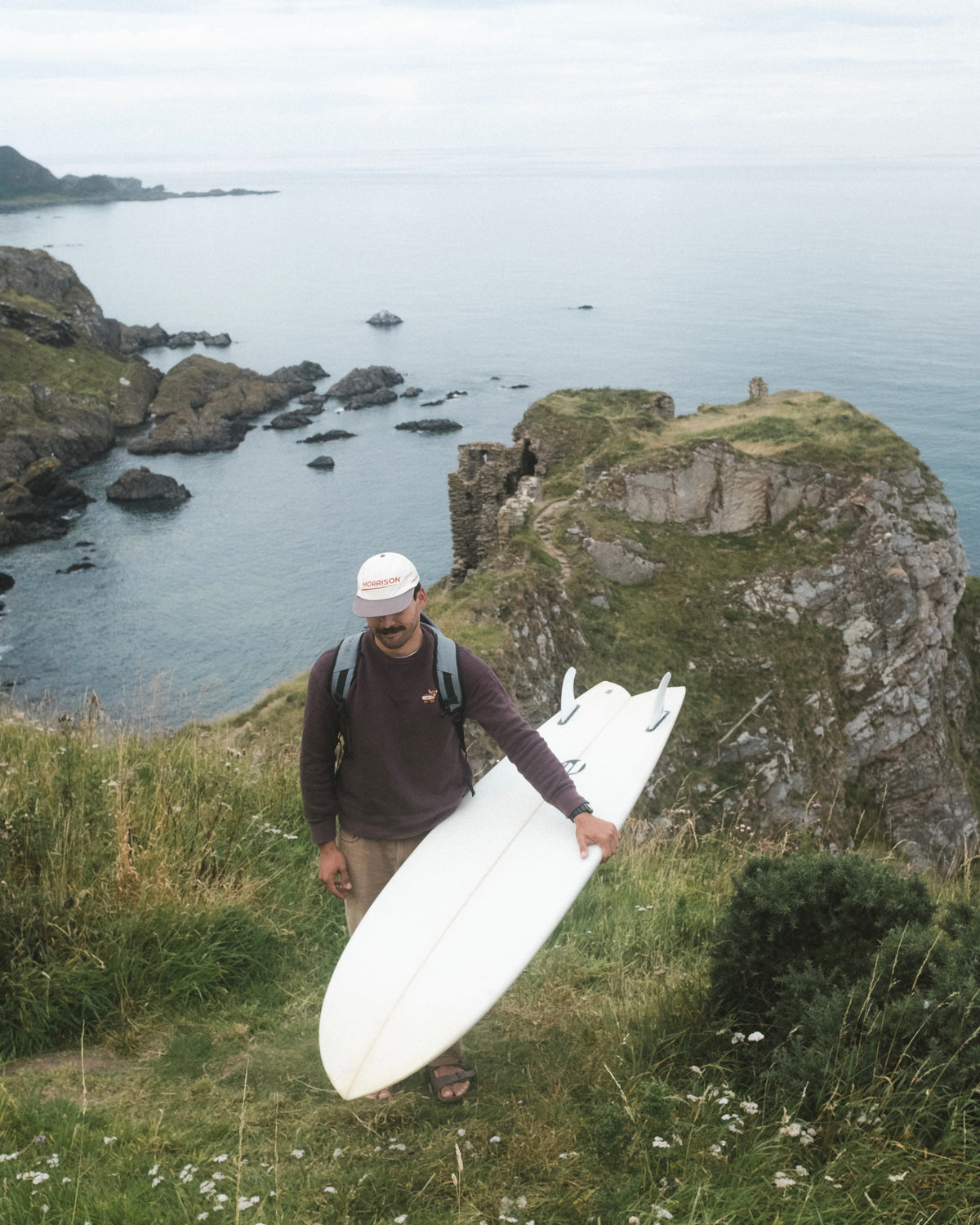 A man carrying a white surfboard down a grassy cliffside overlooking the ocean, wearing a dark brown long-sleeve shirt, beige pants, and a white cap.
